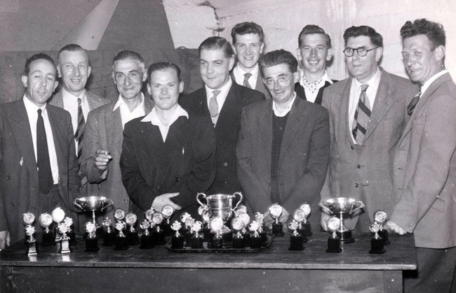 The Royal Oak Skittles Team stand behind a table laden with trophies.